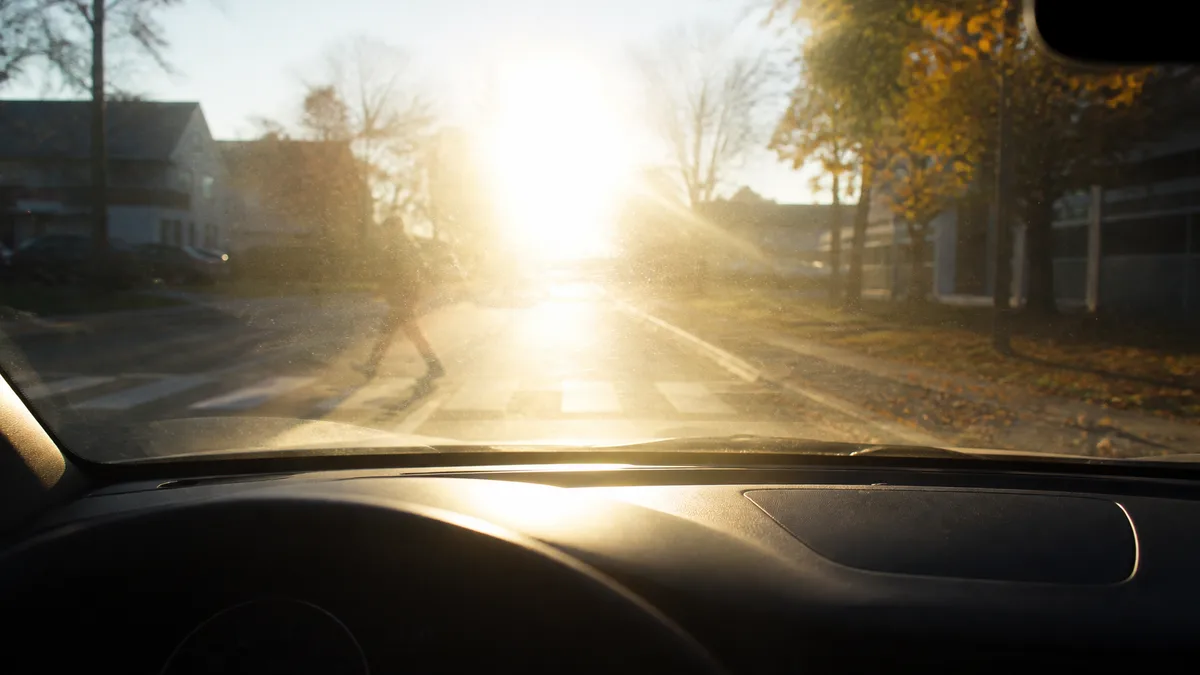 Driver's-seat view through a windshield into low, glaring morning sun on a residential street near a school zone crosswalk